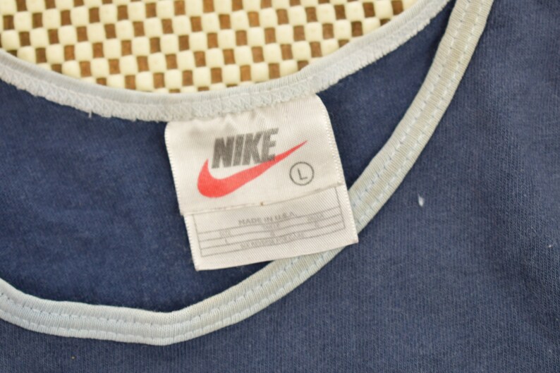 May include: A navy blue tank top with a white trim around the neckline. The tag on the shirt says "NIKE" in white letters with a red swoosh logo and "MADE IN U.S.A." in black text. The tag also has a size "L" in a white circle.