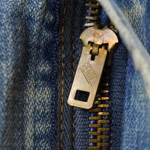 Puede incluir: Primer plano de una cremallera de lat&oacute;n sobre tela vaquera azul. El tirador de la cremallera es rectangular con la palabra "PRENTICE" grabada. Los dientes de la cremallera son dorados.