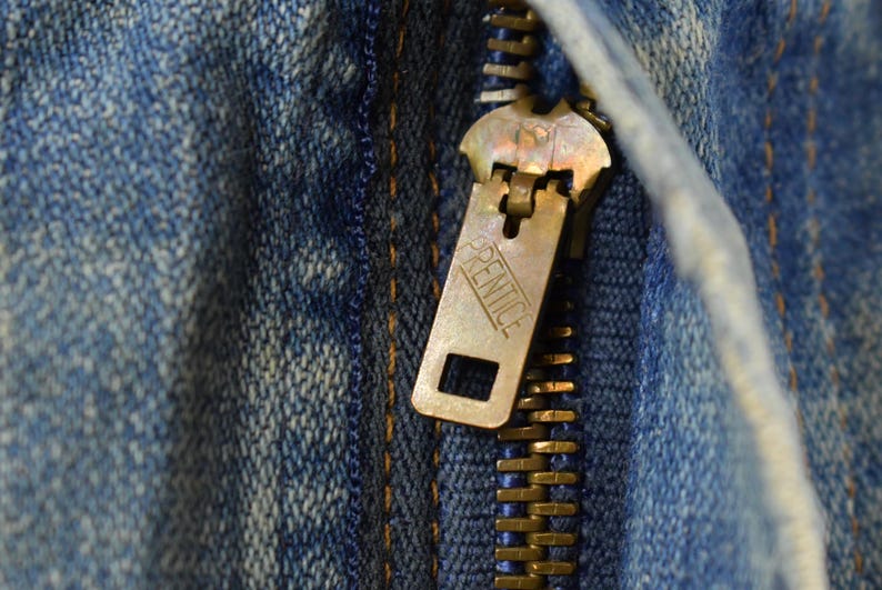 Puede incluir: Primer plano de una cremallera de lat&oacute;n sobre tela vaquera azul. El tirador de la cremallera es rectangular con la palabra "PRENTICE" grabada. Los dientes de la cremallera son dorados.