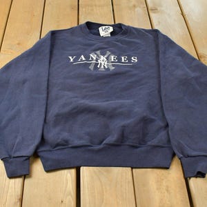 May include: A navy blue long-sleeve crewneck sweatshirt featuring the word YANKEES in white lettering across the chest, with the interlocking NY logo centred below the text. The garment includes a Lee brand tag at the inner neckline.