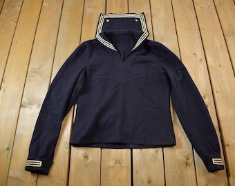 May include: A navy blue long-sleeved shirt with a white sailor collar and white trim on the cuffs. The shirt has a v-neck and a pocket on the chest.