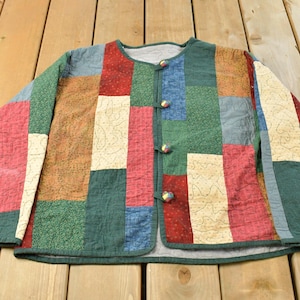 Puede incluir: Una chaqueta acolchada de manga larga con un diseño de patchwork de paneles rectangulares de tela en tonos rojo, verde, azul y crema. La chaqueta tiene un cuello redondo, un cierre frontal con cuatro botones cubiertos de tela y costuras.