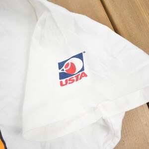 Puede incluir: Camiseta blanca con un logotipo de tenis azul y rojo que dice "USTA" en la manga derecha. La camiseta tambi&eacute;n tiene un gr&aacute;fico de dibujos animados en la parte delantera.