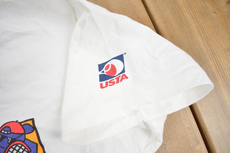 Puede incluir: Camiseta blanca con un logotipo de tenis azul y rojo que dice "USTA" en la manga derecha. La camiseta tambi&eacute;n tiene un gr&aacute;fico de dibujos animados en la parte delantera.