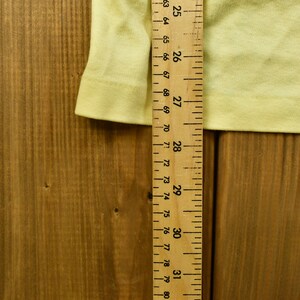 K&ouml;nnte beinhalten: Nahaufnahme eines gelben Stoffes mit einem Holzlineal, das 78,7 cm misst. Das Lineal ist mit Zahlen von 25 bis 31 gekennzeichnet.