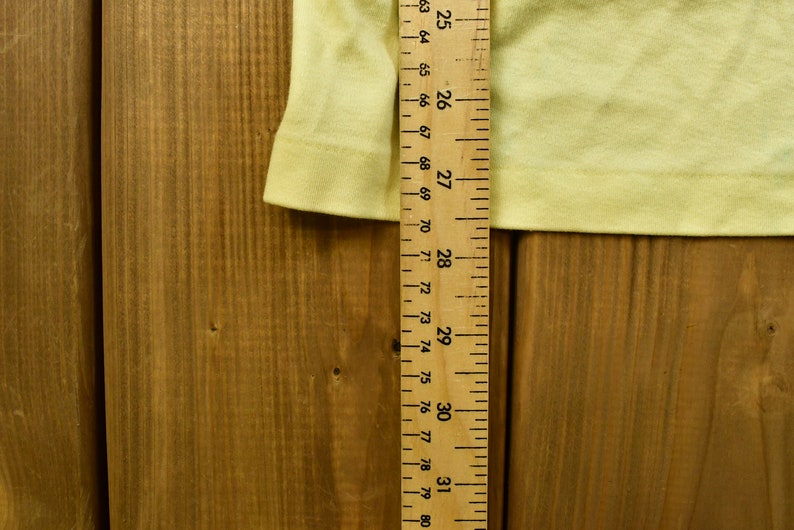 K&ouml;nnte beinhalten: Nahaufnahme eines gelben Stoffes mit einem Holzlineal, das 78,7 cm misst. Das Lineal ist mit Zahlen von 25 bis 31 gekennzeichnet.