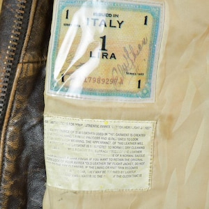 May include: Close-up of a brown leather jacket interior with a zipper and a label that reads "ISSUED IN ITALY". The label also includes the text "1 LIRA" and a serial number. The jacket's lining is a light beige color.
