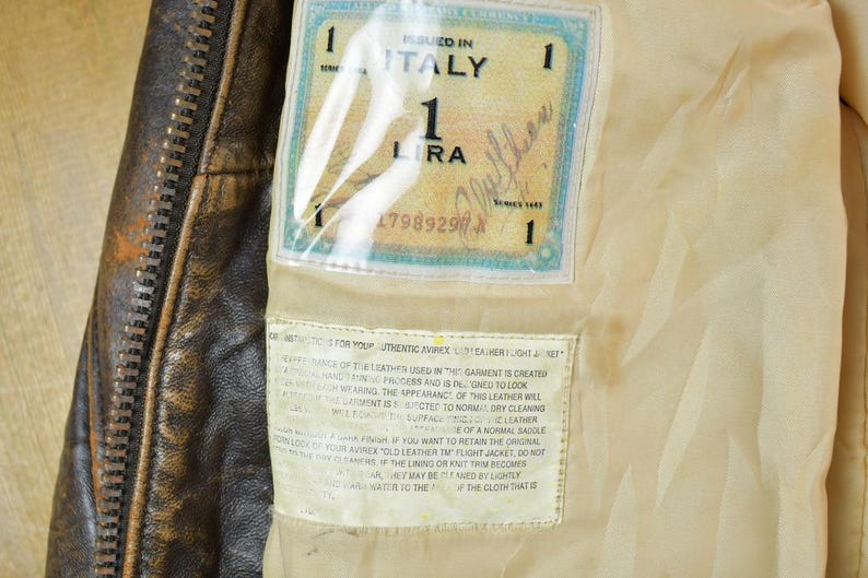 May include: Close-up of a brown leather jacket interior with a zipper and a label that reads "ISSUED IN ITALY". The label also includes the text "1 LIRA" and a serial number. The jacket's lining is a light beige color.