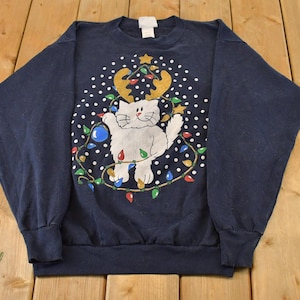 Puede incluir: Una sudadera azul marino de manga larga con un gráfico de un gato blanco que lleva astas de reno doradas. El gato está rodeado por una cadena de luces navideñas de colores y puntos blancos sobre un fondo azul oscuro.