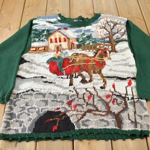 Peut inclure: Un pull en tricot vert avec une scène d'hiver représentant un traîneau tiré par un cheval, une maison et un paysage enneigé. Le pull a une bordure verte et un fond blanc.