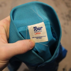 May include: A close-up of a teal jacket collar with a white tag. The tag reads "Tour MADE IN U.S.A. LARGE CARE INSTRUCTIONS OVER RN 71662". The jacket is teal with a ribbed knit collar.