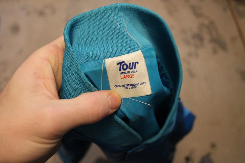 May include: A close-up of a teal jacket collar with a white tag. The tag reads "Tour MADE IN U.S.A. LARGE CARE INSTRUCTIONS OVER RN 71662". The jacket is teal with a ribbed knit collar.