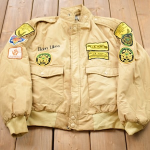 May include: Tan bomber jacket with multiple patches, including "Eldon Likes" text and "J.B. Hunt" patches. Features a button-up front, two front pockets, and ribbed cuffs and hem. The jacket has a classic, vintage style.