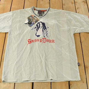 Può includere: T-shirt grigio chiaro con grafica Snoop Dogg. La maglietta ha uno scollo a V, finiture blu navy e il logo Snoop Dogg in rosso. Un'etichetta è attaccata vicino al colletto. La maglietta è appoggiata su una superficie di legno.