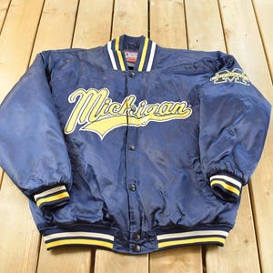 May include: A navy blue varsity jacket featuring the word Michigan in yellow script across the chest. The jacket has a snap-button front closure, a ribbed collar, cuffs, and hem with yellow and white stripes, and a Michigan logo on the left sleeve.