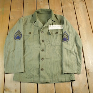 Könnte beinhalten: Olivgrüne Militärjacke mit Knopfleiste, zwei Brusttaschen und Aufnähern auf den Ärmeln. Die Aufnäher zeigen blaue und goldene Winkel. Ein weißes Namensschild befindet sich über der rechten Brusttasche.