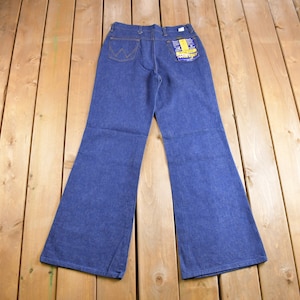 Op de afbeelding: Een paar blauwe denim bell-bottom jeans met een geel en blauw label op de achterzak.