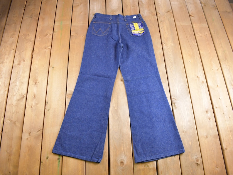 Op de afbeelding: Een paar blauwe denim bell-bottom jeans met een geel en blauw label op de achterzak.