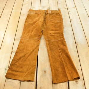 May include: A pair of tan suede leather pants featuring a straight-leg cut, a front button closure, two large patch pockets on the front, and horizontal seam detailing across the mid-thigh area of both legs, presented in a flat, full-length view.