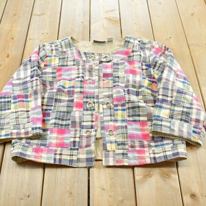 May include: A cropped jacket featuring a multi-coloured patchwork plaid pattern in shades of pink, blue, yellow, and white. The jacket has three-quarter length sleeves, a round neckline, and a front closure with four silver-toned metal grommets.