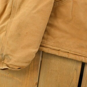 Puede incluir: Primer plano de una chaqueta de trabajo color beige, que muestra la manga y parte del cuerpo. La chaqueta tiene un forro acolchado y un bolsillo. La tela es de lona resistente, con algo de desgaste visible.