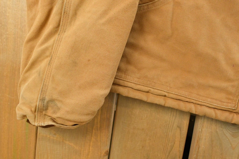 Puede incluir: Primer plano de una chaqueta de trabajo color beige, que muestra la manga y parte del cuerpo. La chaqueta tiene un forro acolchado y un bolsillo. La tela es de lona resistente, con algo de desgaste visible.