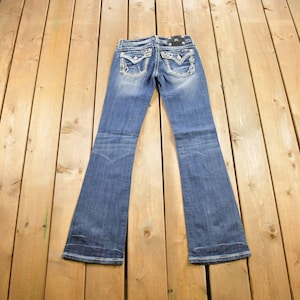 Peut inclure: Un jean bleu foncé en denim avec une coupe pattes d'éléphant. Le jean a un ornement en strass sur les poches arrière.