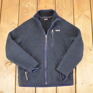 May include: A navy blue fleece jacket with a full zipper closure, two side pockets, and a stand-up collar. The jacket features a small logo patch on the chest and a navy blue zipper. The jacket is made of a soft, textured material.
