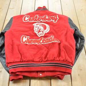 May include: A red and black letterman jacket with the words "Centerburg Cheerleaders" in white script. The jacket has black leather sleeves and a red body. The jacket has a white emblem of a helmeted figure.