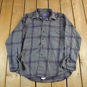 Puede incluir: Camisa abotonada a cuadros grises y morados. La camisa de manga larga tiene dos bolsillos en el pecho y un cuello abotonado. La camisa est&aacute; hecha de una tela suave y tejida y est&aacute; colocada sobre una superficie de madera.