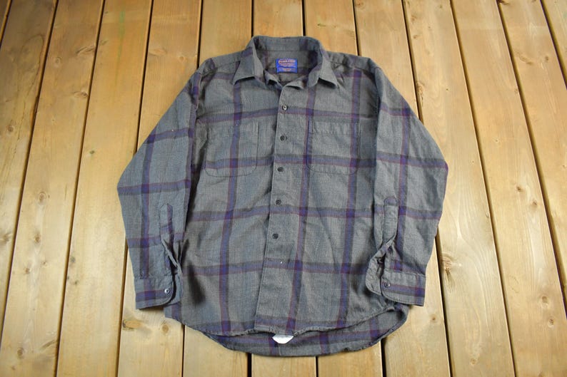 Puede incluir: Camisa abotonada a cuadros grises y morados. La camisa de manga larga tiene dos bolsillos en el pecho y un cuello abotonado. La camisa est&aacute; hecha de una tela suave y tejida y est&aacute; colocada sobre una superficie de madera.