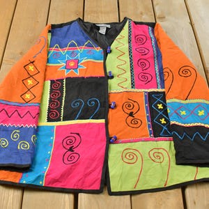 May include: A long-sleeve jacket featuring a vibrant patchwork design with blocks of orange, blue, pink, black, and lime green. The garment is decorated with embroidered patterns including swirls, zigzags, and stars, and fastens with three blue toggles.