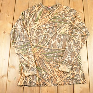 May include: A long-sleeved camouflage shirt with a crew neck. The shirt features a pattern of brown, tan, and green, resembling a woodland or marsh environment. The shirt is made of a soft material and is designed for outdoor activities.
