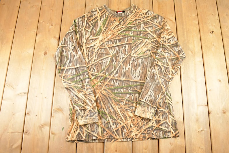 May include: A long-sleeved camouflage shirt with a crew neck. The shirt features a pattern of brown, tan, and green, resembling a woodland or marsh environment. The shirt is made of a soft material and is designed for outdoor activities.
