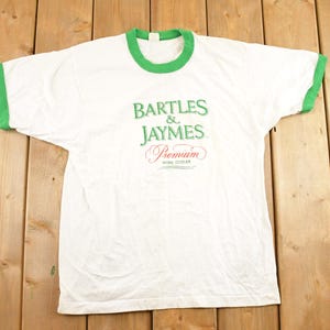 Op de afbeelding: Wit T-shirt met groene randen op de kraag en mouwen. Het shirt heeft de tekst "BARTLES & JAYMES Premium Wine Cooler" in groene en rode letters. Het shirt is gemaakt van een zacht materiaal.
