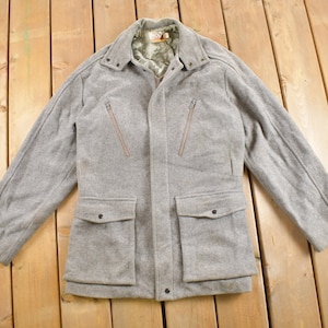 May include: A light grey wool jacket with a collared neckline, two chest pockets, and two lower pockets. The jacket has a zip closure and a quilted lining. The jacket is laid flat on a wooden surface.