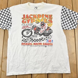 Puede incluir: Camiseta blanca con un patrón a cuadros en las mangas. La parte delantera de la camiseta presenta un gráfico de un piloto de motocicleta con el texto "Jackpine Gypsies Classic Motorcycle Racing 1997 Pro-Am Racing Sturgis, South Dakota AMA"
