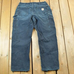 May include: Dark blue work pants with multiple pockets and a small logo patch. The pants are made of a durable material and have a relaxed fit. The work pants are laid out on a wooden surface.