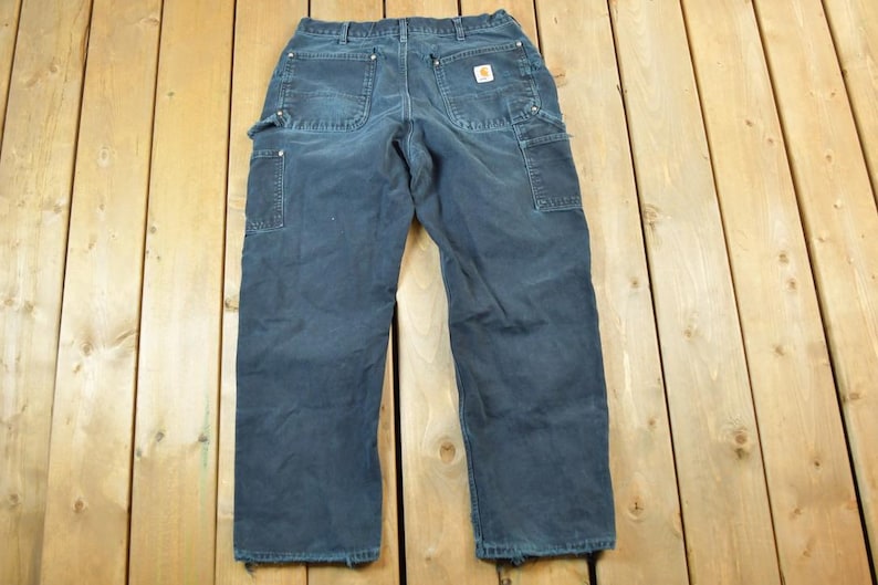 May include: Dark blue work pants with multiple pockets and a small logo patch. The pants are made of a durable material and have a relaxed fit. The work pants are laid out on a wooden surface.