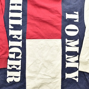 Pode incluir: Uma camisa de manga comprida com um design de blocos de cores vermelho, branco e azul. A camisa apresenta as palavras "Tommy" e "Hilfiger" em letras brancas nas mangas.