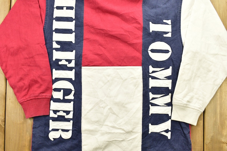 Pode incluir: Uma camisa de manga comprida com um design de blocos de cores vermelho, branco e azul. A camisa apresenta as palavras "Tommy" e "Hilfiger" em letras brancas nas mangas.