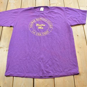 May include: A purple t-shirt with a crew neck and short sleeves. The shirt features a circular graphic with the text "Galway Rowing Clubs Boston Ma 1st USA Visit" in a gold color. The shirt is laid flat on a wooden surface.