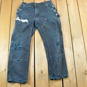 May include: Dark blue work pants with distressed details, including ripped knees and frayed edges. The pants have multiple pockets and white accents. They are laid flat on a wooden surface.