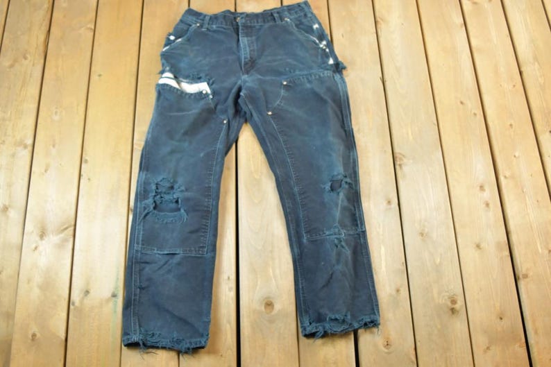 May include: Dark blue work pants with distressed details, including ripped knees and frayed edges. The pants have multiple pockets and white accents. They are laid flat on a wooden surface.