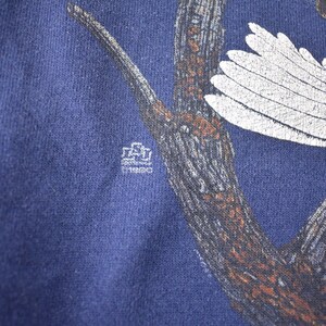 K&ouml;nnte beinhalten: Ein marineblaues T-Shirt mit einem wei&szlig;en Adler, der auf einem braunen Ast sitzt. Der Adler hat einen wei&szlig;en Fl&uuml;gel ausgebreitet. Das T-Shirt hat ein kleines wei&szlig;es Etikett mit dem Text "Southwestern University" darauf.
