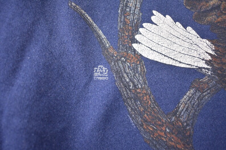 K&ouml;nnte beinhalten: Ein marineblaues T-Shirt mit einem wei&szlig;en Adler, der auf einem braunen Ast sitzt. Der Adler hat einen wei&szlig;en Fl&uuml;gel ausgebreitet. Das T-Shirt hat ein kleines wei&szlig;es Etikett mit dem Text "Southwestern University" darauf.