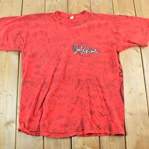 May include: Red t-shirt with a black pattern and the "Body Glove" logo in white script. The shirt has short sleeves and a crew neck. The fabric appears to be slightly faded.