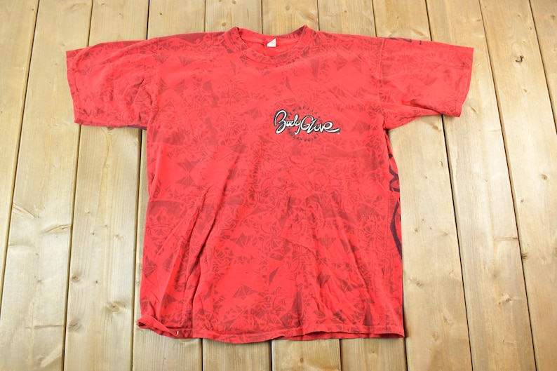 May include: Red t-shirt with a black pattern and the "Body Glove" logo in white script. The shirt has short sleeves and a crew neck. The fabric appears to be slightly faded.