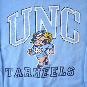 May include: A light blue long-sleeved t-shirt with a white outline of "UNC" and the words "Tarheels" in navy blue. A cartoon mascot with a blue and white hat and a blue and white sweater is in the center of the shirt.
