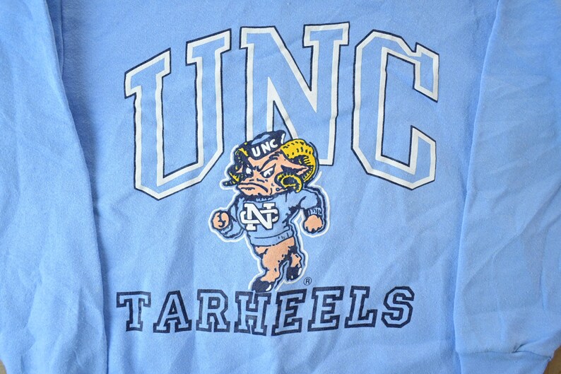 May include: A light blue long-sleeved t-shirt with a white outline of "UNC" and the words "Tarheels" in navy blue. A cartoon mascot with a blue and white hat and a blue and white sweater is in the center of the shirt.
